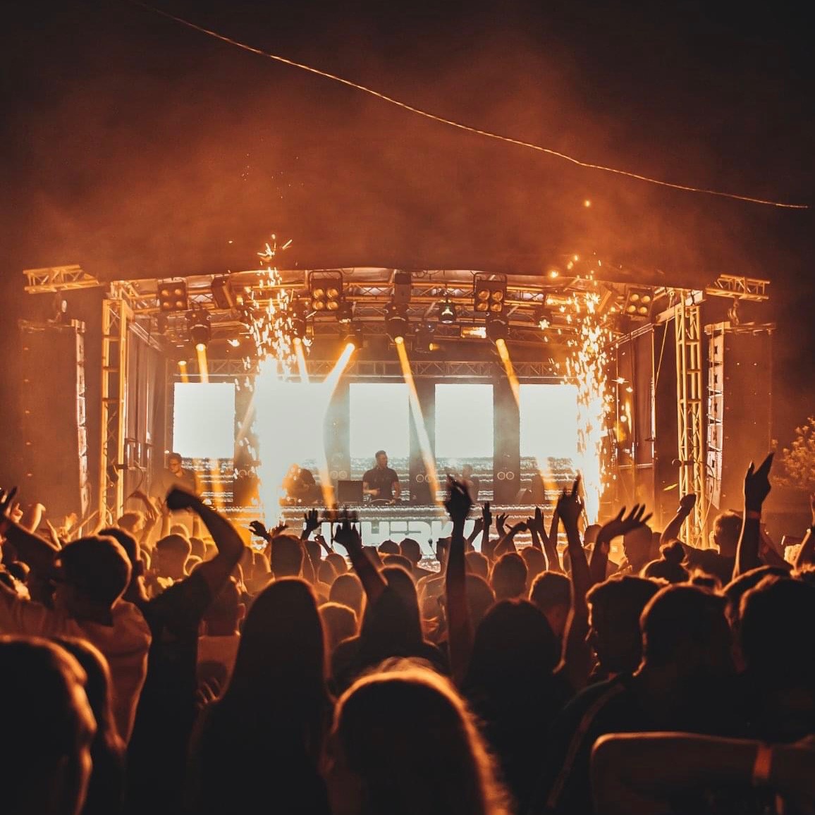 Tłum ludzi z podniesionymi rękami podczas koncertu na zewnątrz, na scenie DJ i efekty pirotechniczne.