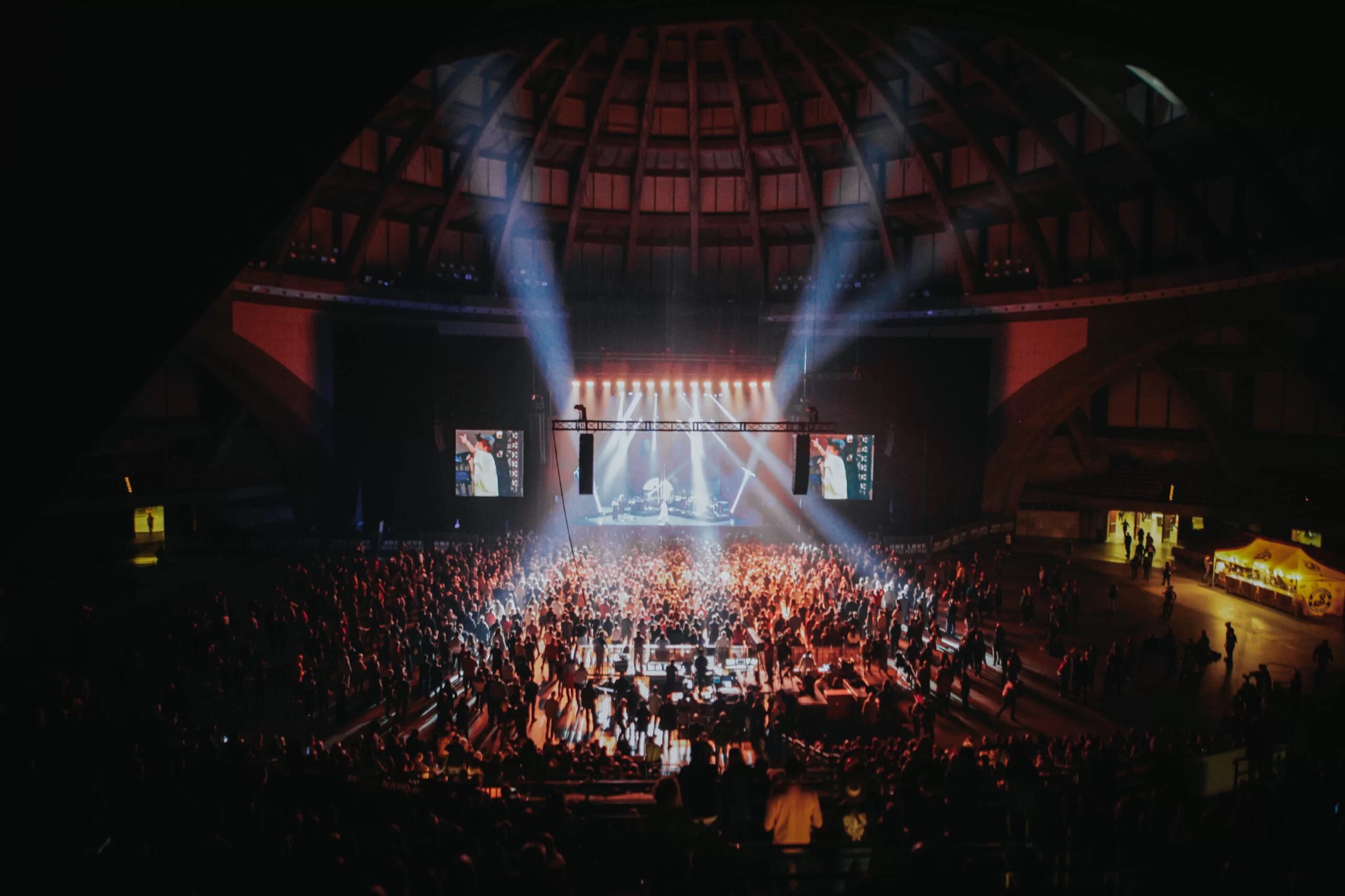 Widok z góry na dużą halę koncertową wypełnioną publicznością, scena oświetlona reflektorami, z widocznymi ekranami po bokach, charakterystyczna kopuła hali w tle.