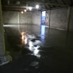 Świeżo wylana, mokra posadzka przemysłowa w surowym, oświetlonym hali. Widoczne refleksy światła na powierzchni wylewki i ściany z betonu kom&oacute;rkowego.