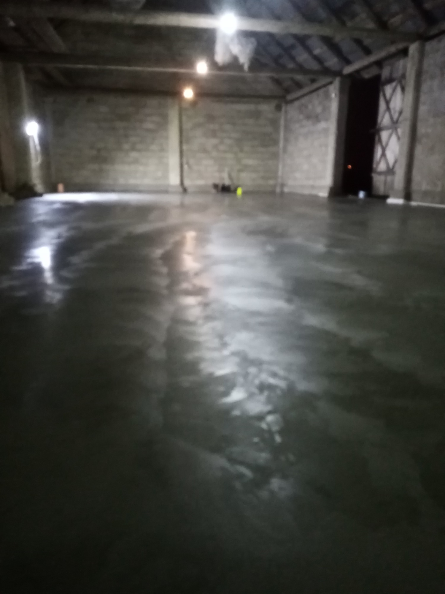 Świeżo wylana, mokra posadzka w hali w Grajewie, z widocznymi śladami po narzędziach i odbiciami światła. W tle surowe, betonowe ściany i drewniana konstrukcja dachu.