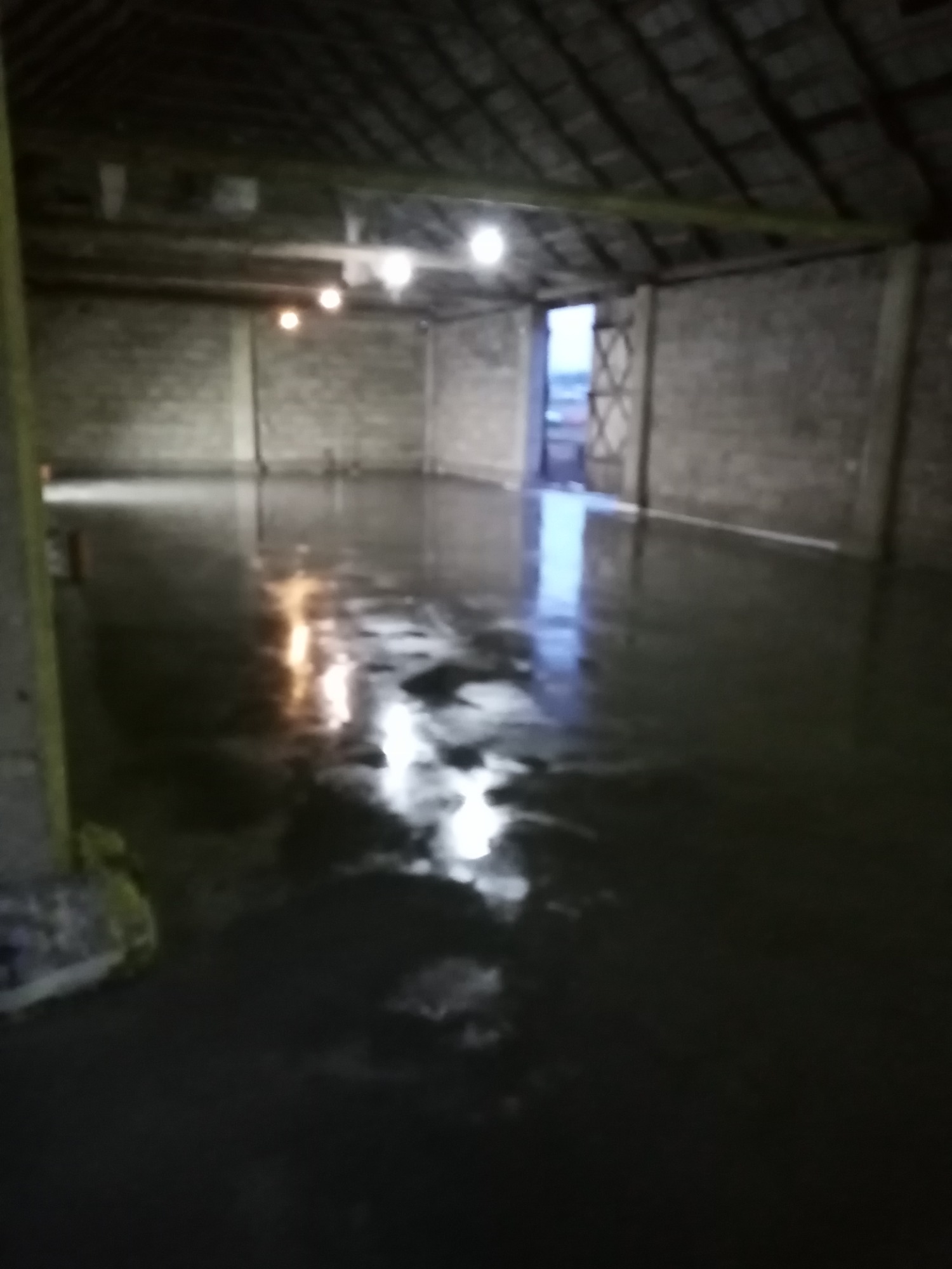 Świeżo wylana, mokra posadzka przemysłowa w surowym, oświetlonym hali. Widoczne refleksy światła na powierzchni wylewki i ściany z betonu komórkowego.