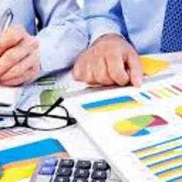 Dwie osoby analizują kolorowe wykresy i dokumenty, używając długopisów, z kalkulatorami, okularami i laptopem na biurku.