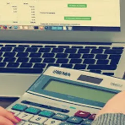 Osoba używa kalkulatora obok otwartego laptopa z arkuszem kalkulacyjnym na drewnianym biurku, widoczny segregator i szklanka wody.