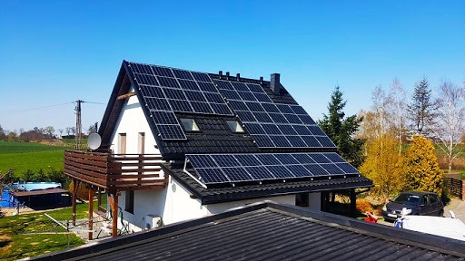 Dom jednorodzinny z zainstalowanymi panelami słonecznymi na dachu, widok z góry, w tle zielone pola i błękitne niebo.