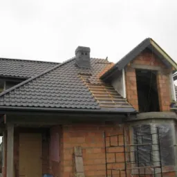 Dom w trakcie budowy z częściowo pokrytym dachem z ciemnej blachodachówki i widoczną konstrukcją więźby dachowej na lukarnie, komin wykończony w podobnej kolorystyce.