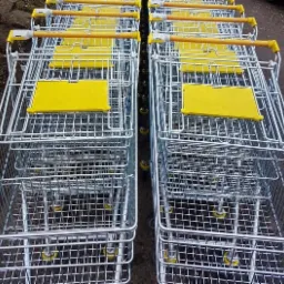 Rząd metalowych wózków sklepowych z żółtymi elementami, ustawionych jeden za drugim na zewnątrz.