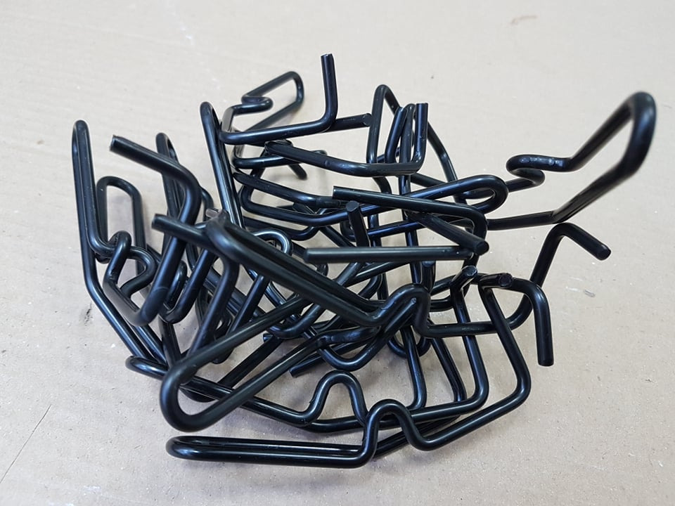 Stos czarnych, metalowych, giętych elementów o nieregularnym kształcie, prawdopodobnie uchwytów lub mocowań, na neutralnym tle.