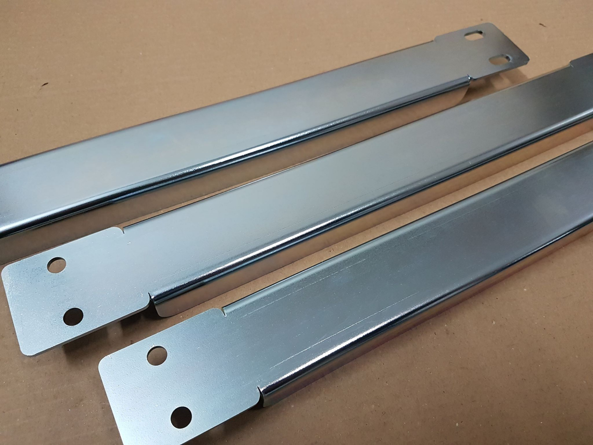 Trzy metalowe, prostokątne profile z otworami, prawdopodobnie stalowe, z błyszczącą, srebrną powłoką, leżące na tekturowym podłożu.