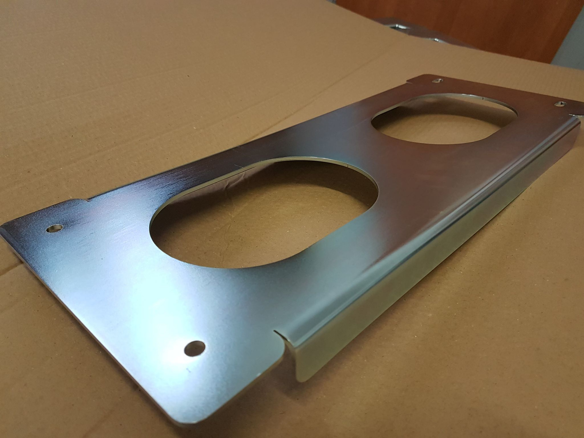 Metalowa płyta z dwoma okrągłymi otworami i otworami montażowymi, leżąca na kartonowym podłożu, prezentująca gładką, jednolitą powierzchnię.