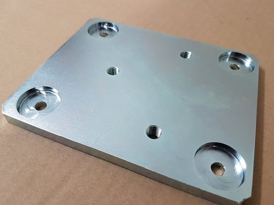 Szlifowana, srebrna, kwadratowa płyta metalowa z otworami o różnej średnicy i gwintowanymi otworami, leżąca na brązowym tle.