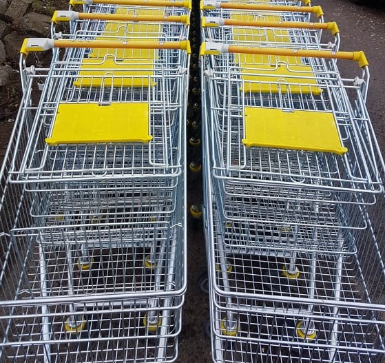 Rząd metalowych wózków sklepowych z żółtymi elementami, ustawionych jeden za drugim na zewnątrz.