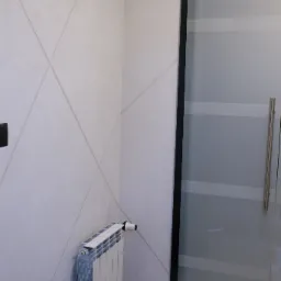 Ściana z dekoracyjną, jasnoszarą strukturą i geometrycznymi liniami, biały grzejnik panelowy pod ścianą, przesuwna szklana ścianka działowa z poziomymi pasami.