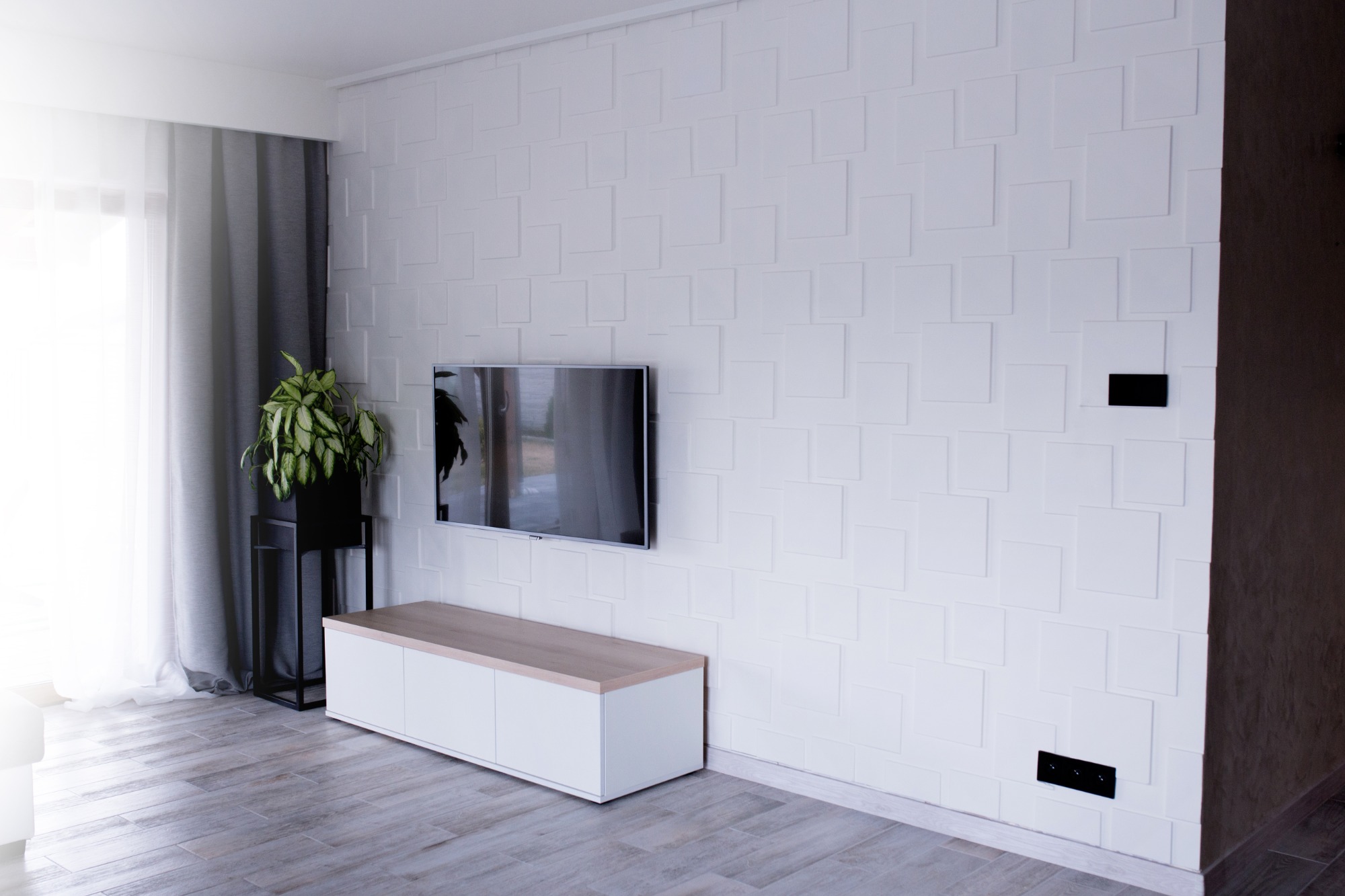 Nowoczesny salon z białą ścianą 3D, telewizorem, szafką RTV i rośliną doniczkową w czarnym stojaku.