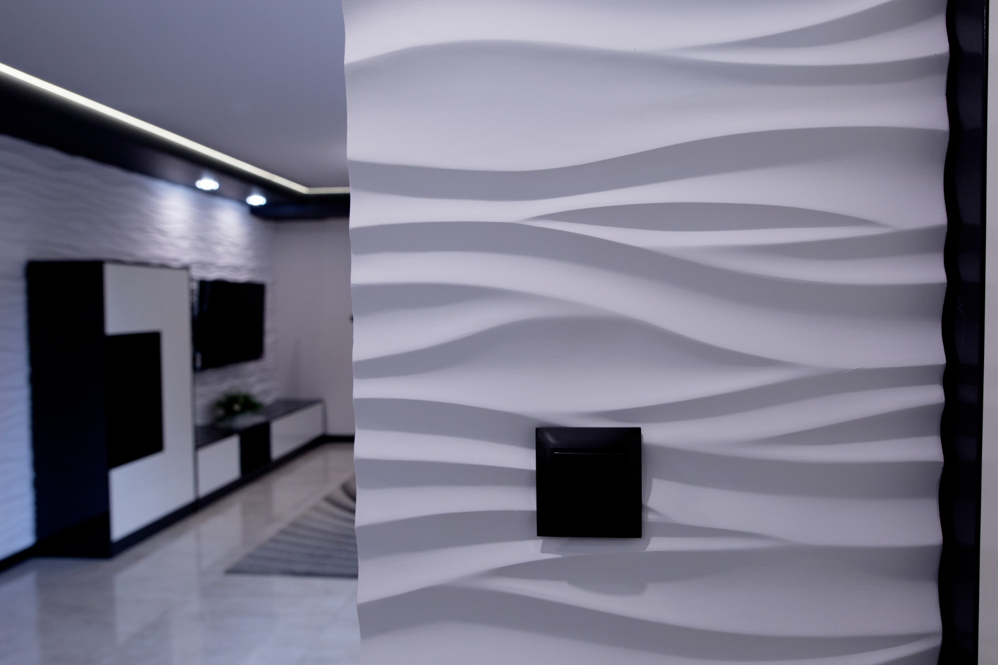 Biała ściana z dekoracyjnymi panelami 3D w kształcie fal, z czarnym włącznikiem światła, w tle widoczny fragment salonu z nowoczesnymi meblami w czarno-białej kolorystyce.