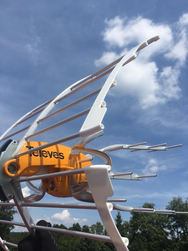 Antena telewizyjna marki Televes z pomarańczowym korpusem na tle błękitnego nieba z chmurami, widok z dołu.