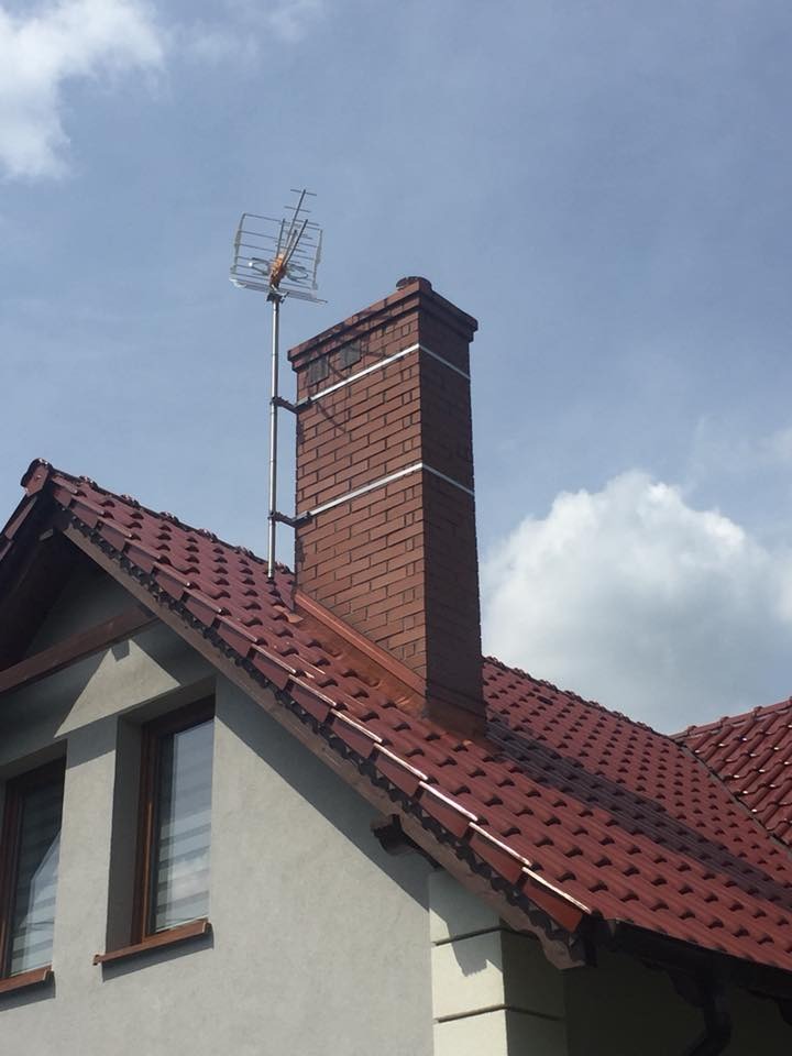 Antena telewizyjna zamontowana na metalowym maszcie przymocowanym do komina z czerwonej cegły na dachu domu z czerwoną dachówką, widok z dołu na błękitne niebo.