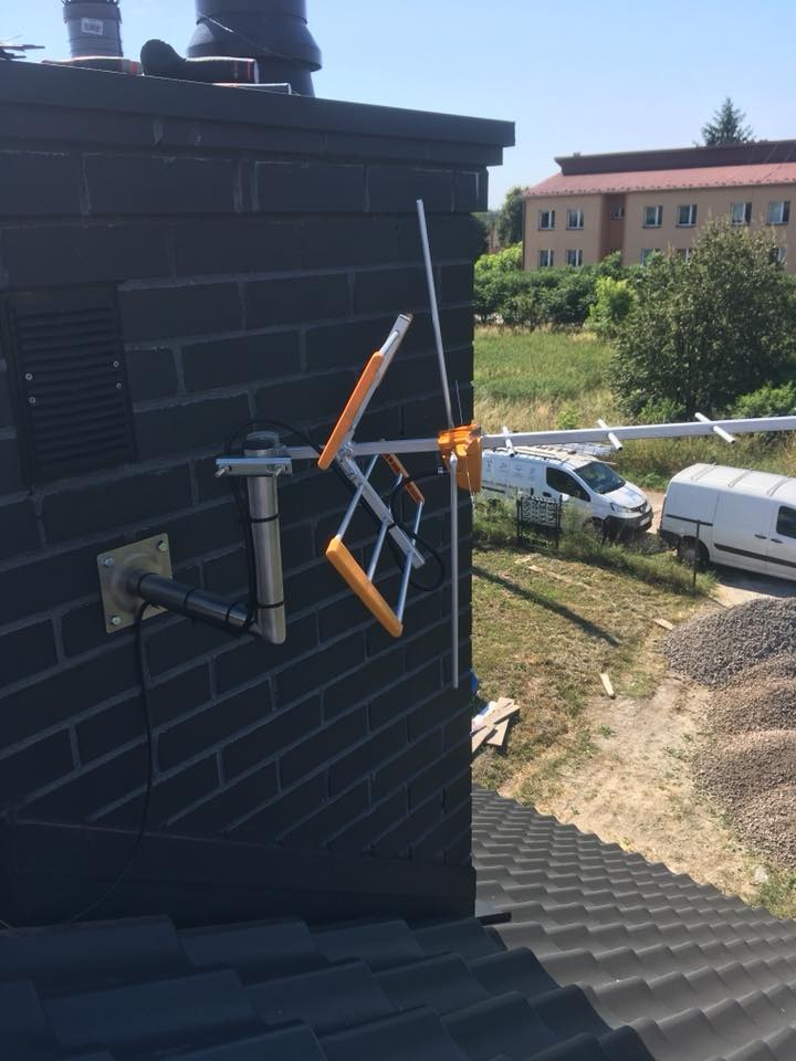 Szara ceglana ściana z zamontowaną anteną telewizyjną z pomarańczowymi elementami, widok z dachu, w tle budynek i zaparkowane białe busy.