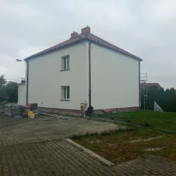 Biały dom po tynkowaniu elewacji; widoczne rusztowania, materiały budowlane i dwie osoby pracujące przy budynku w pochmurny dzień.