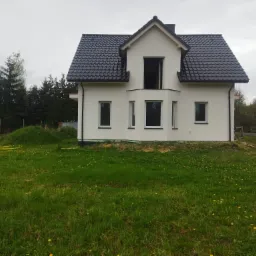 Nowoczesny dom z białymi ścianami i czarnym dachem, otoczony zielonym trawnikiem z żółtymi kwiatami. Widoczne okna i brakujące drzwi balkonowe. Pochmurne niebo.