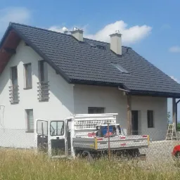 Dom jednorodzinny z nowym, ciemnoszarym dachem z blachodachówki, widoczny zza otwartych drzwi białego samochodu dostawczego na tle wysokiej trawy i fragmentu ogrodzenia z siatki.