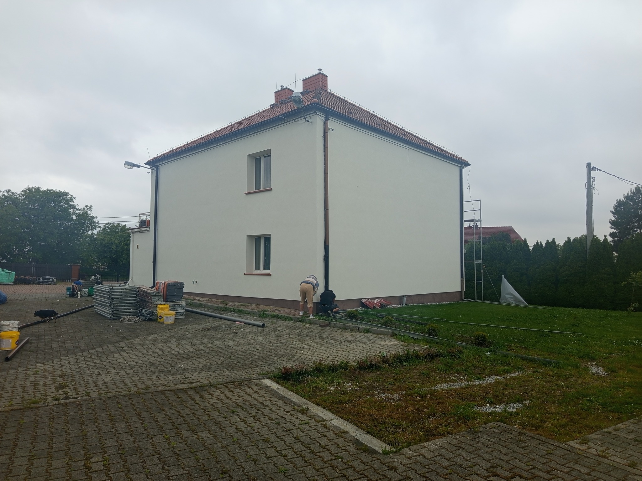 Biały dom po tynkowaniu elewacji; widoczne rusztowania, materiały budowlane i dwie osoby pracujące przy budynku w pochmurny dzień.