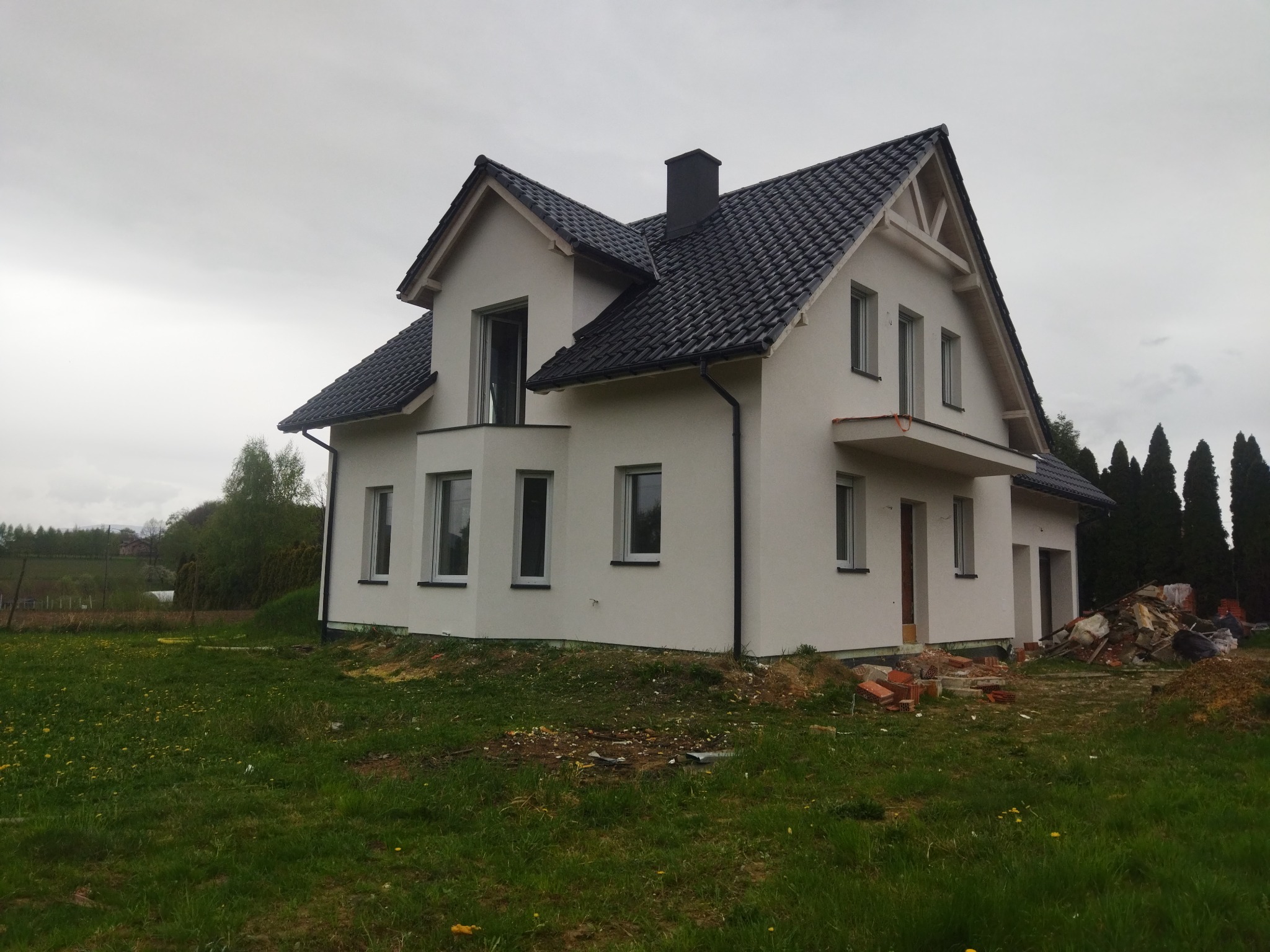 Nowoczesny, dwukondygnacyjny dom z ciemną dachówką i jasną elewacją. Widoczne okna z białymi ramami i fragment terenu z zieloną trawą.