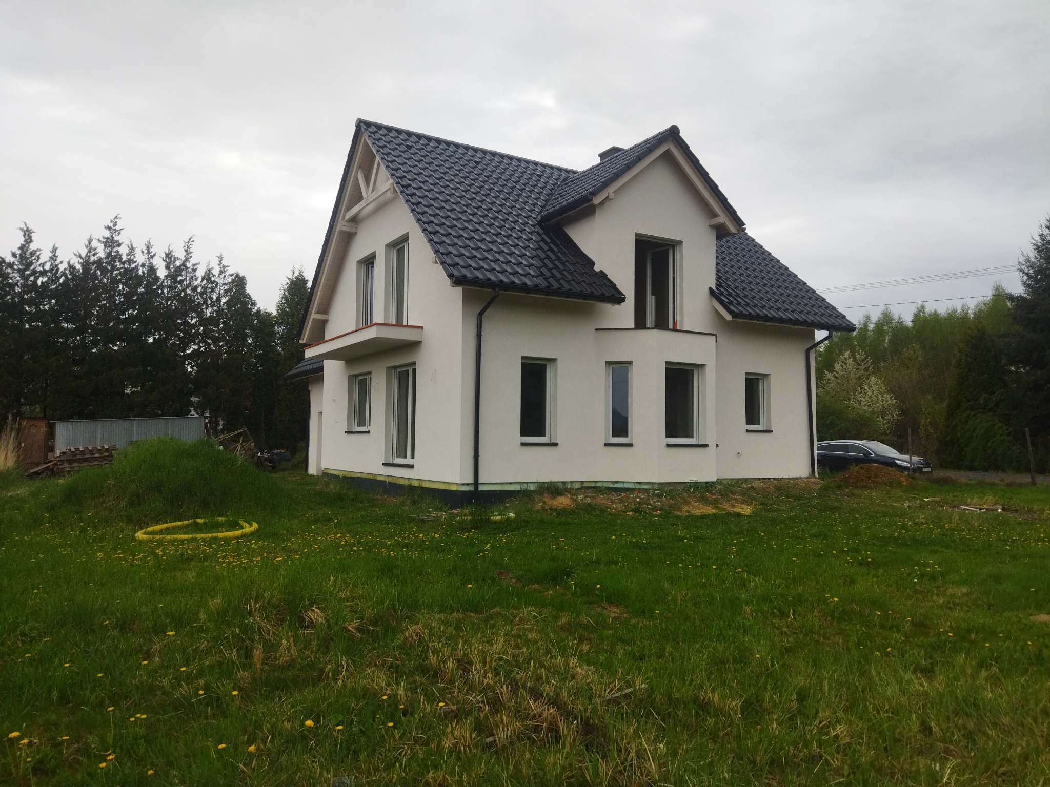 Nowoczesny, dwukondygnacyjny dom z czarnym dachem i jasną elewacją, z otwartym oknem na piętrze i widoczną żółtą rurą na trawniku. Ujęcie z dołu.