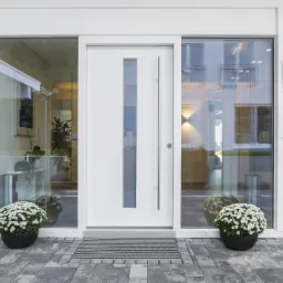 Nowoczesne wejście do domu z białymi drzwiami z przeszkleniem, otoczone dużymi oknami, przed wejściem wycieraczka i dwie donice z białymi kwiatami na szarym bruku.