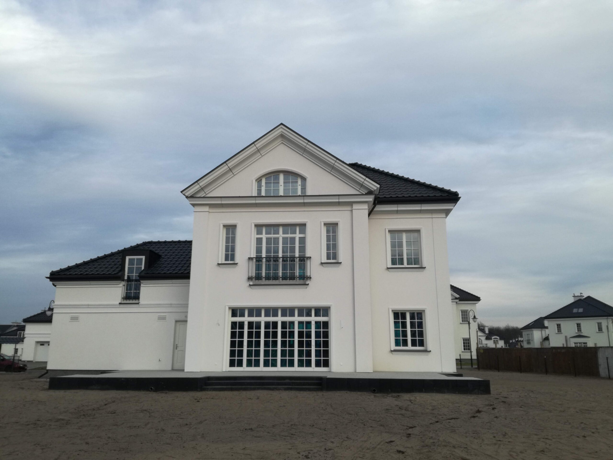 Dwukondygnacyjny dom w stylu klasycystycznym z białymi ścianami, czarnym dachem i dużymi, przeszklonymi drzwiami balkonowymi na parterze oraz oknami z metalową balustradą na piętrze, na tle...