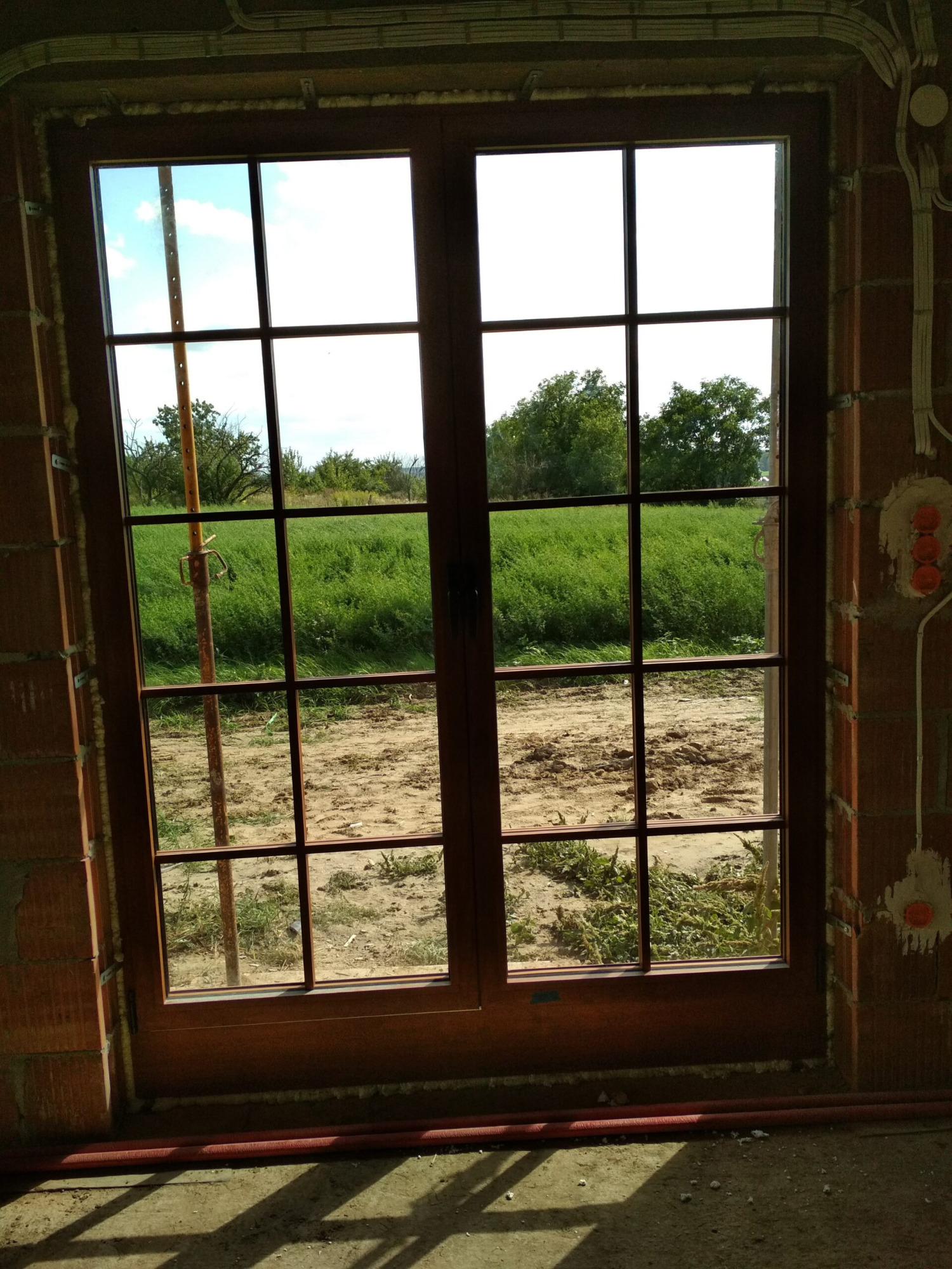 Brązowe okno z widokiem na łąkę, zamontowane w surowym murze z widocznymi instalacjami elektrycznymi i pianką montażową.