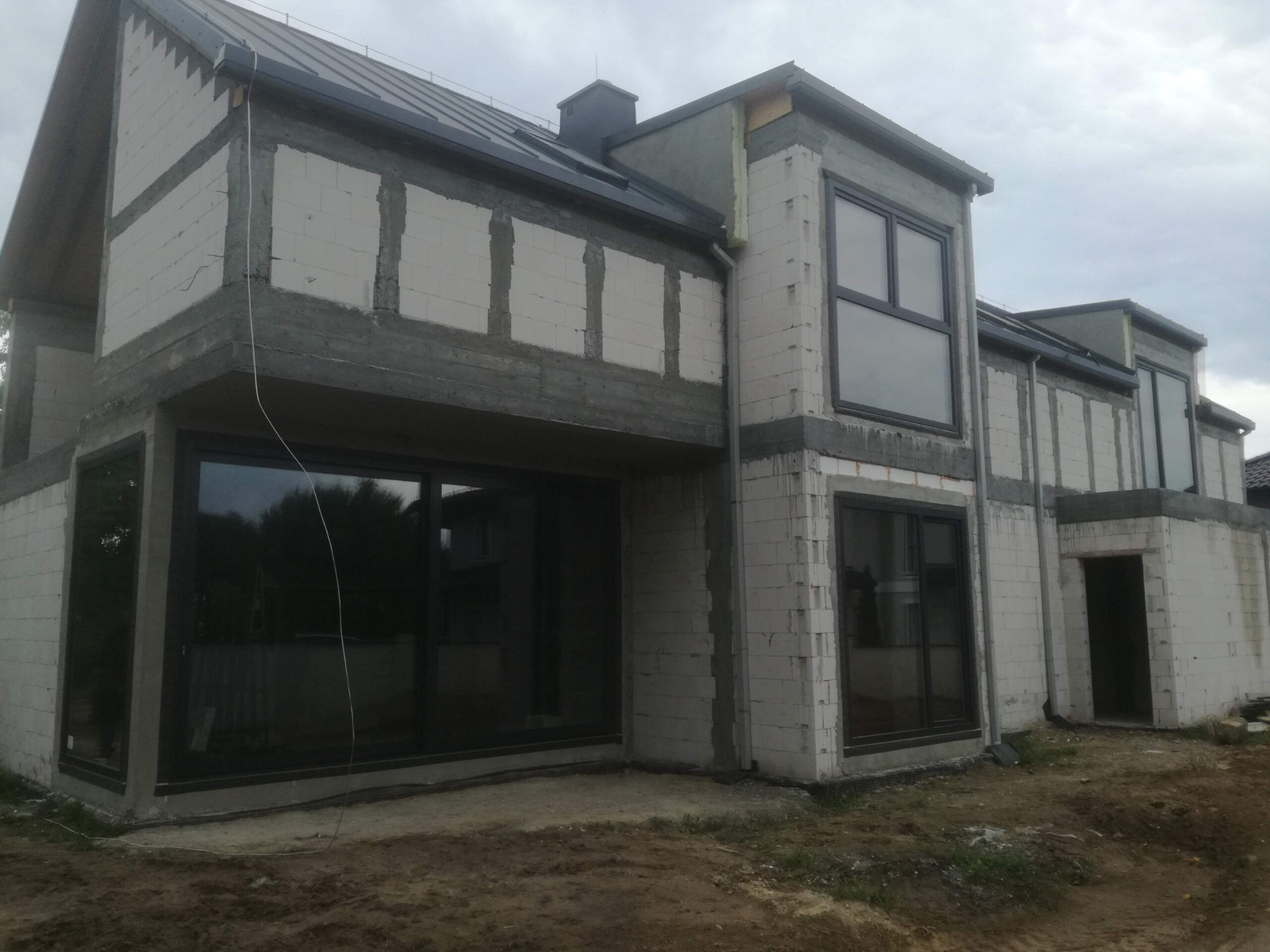 Surowy stan budynku jednorodzinnego z zamontowanymi oknami w czarnych ramach, ściany z betonu komórkowego w trakcie wykończenia, widoczne przewody elektryczne.