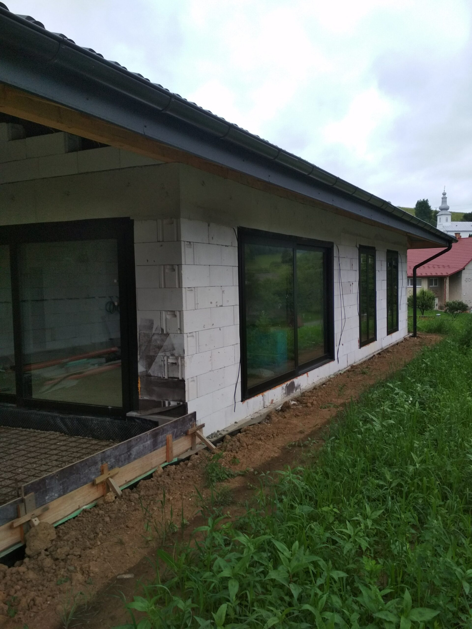Surowy stan budynku z białych bloczków z zamontowanymi ciemnymi oknami, widoczny szalunek fundamentów i trawa, w tle kościół na wzgórzu.