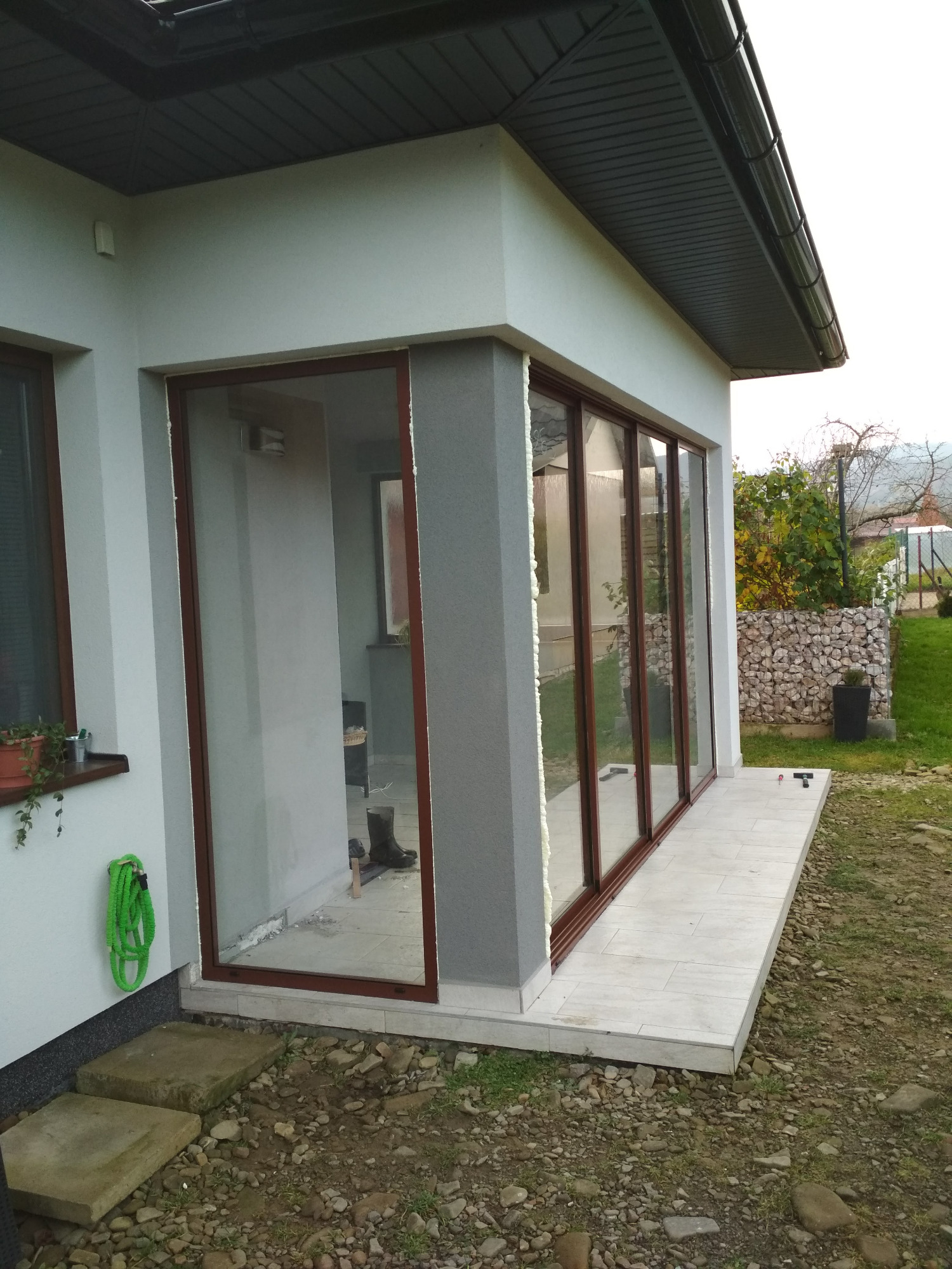 Zewnętrzny widok na dobudowaną werandę z przesuwnymi oknami z brązowymi ramami, widoczne pianka montażowa wokół ram, płytki na podłodze werandy, na ścianie zwinięty zielony wąż ogrodowy.