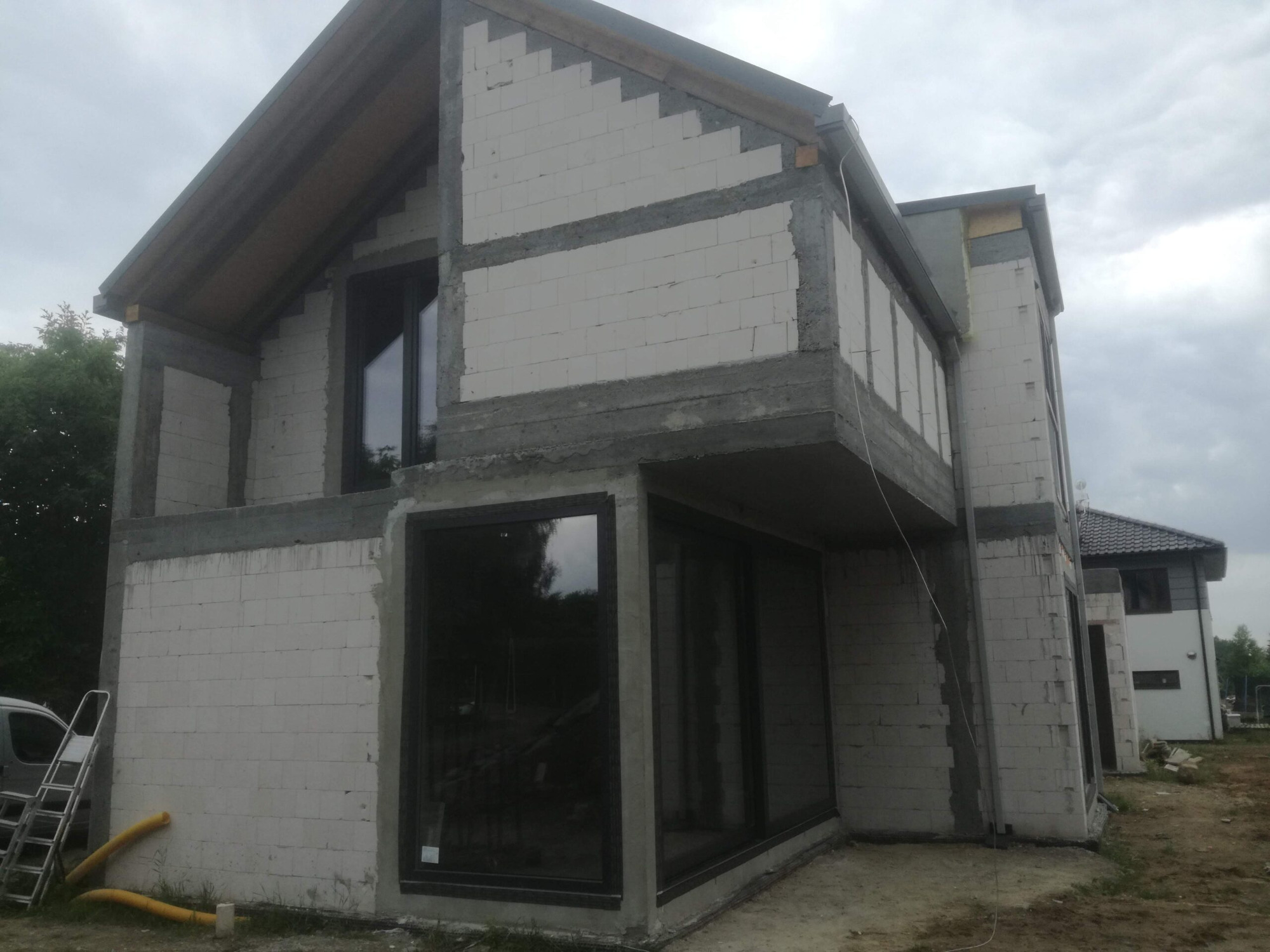 Surowy stan budowy domu jednorodzinnego z zamontowanymi oknami w ciemnych ramach, widoczne białe bloczki, szary beton, drabina oparta o ścianę i żółty wąż na ziemi.