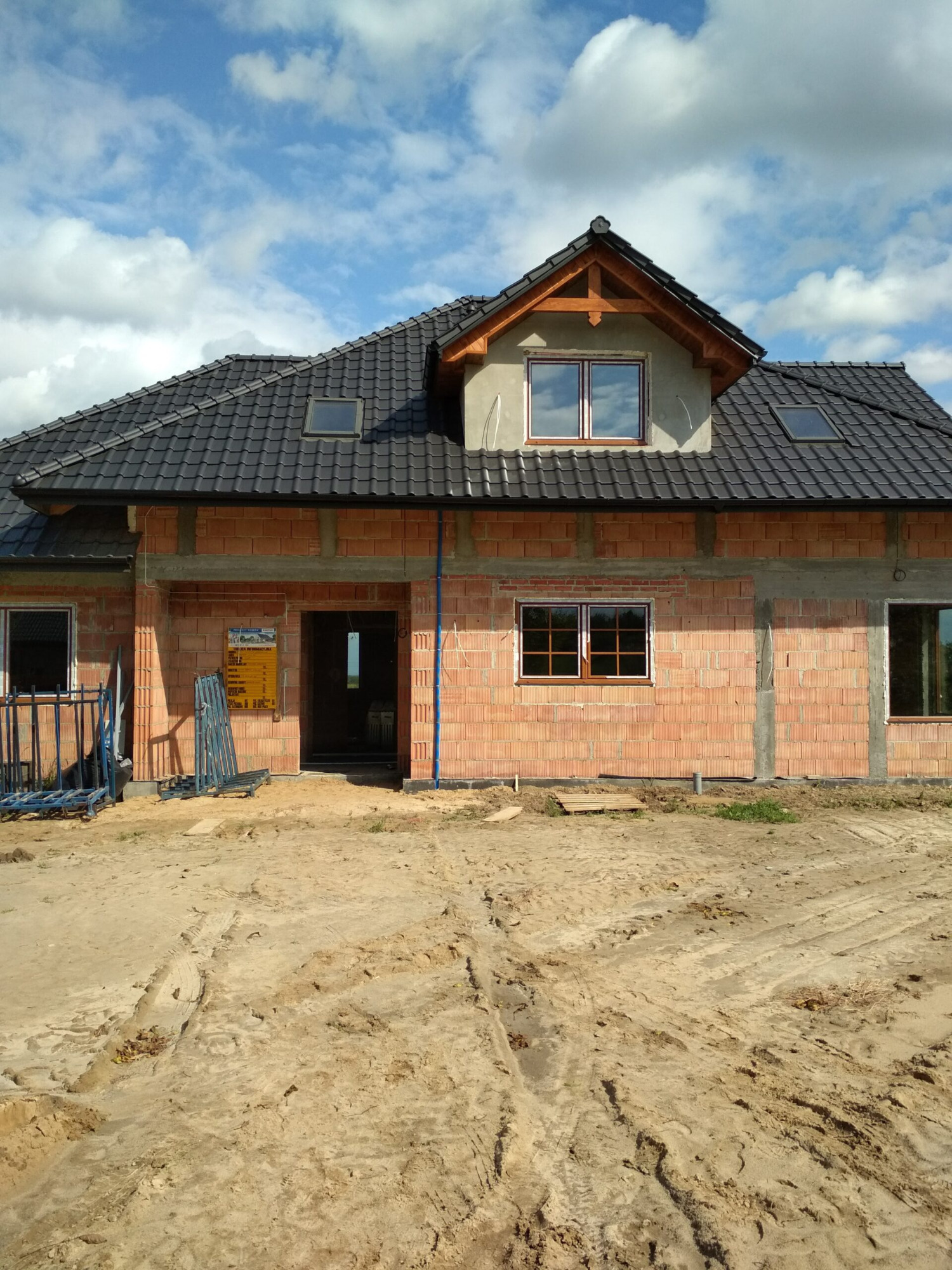 Niedokończony budynek jednorodzinny z czarną dachówką, widoczne okna z brązowymi ramami, ściany z czerwonej cegły, plac budowy z piaskiem i sprzętem budowlanym.