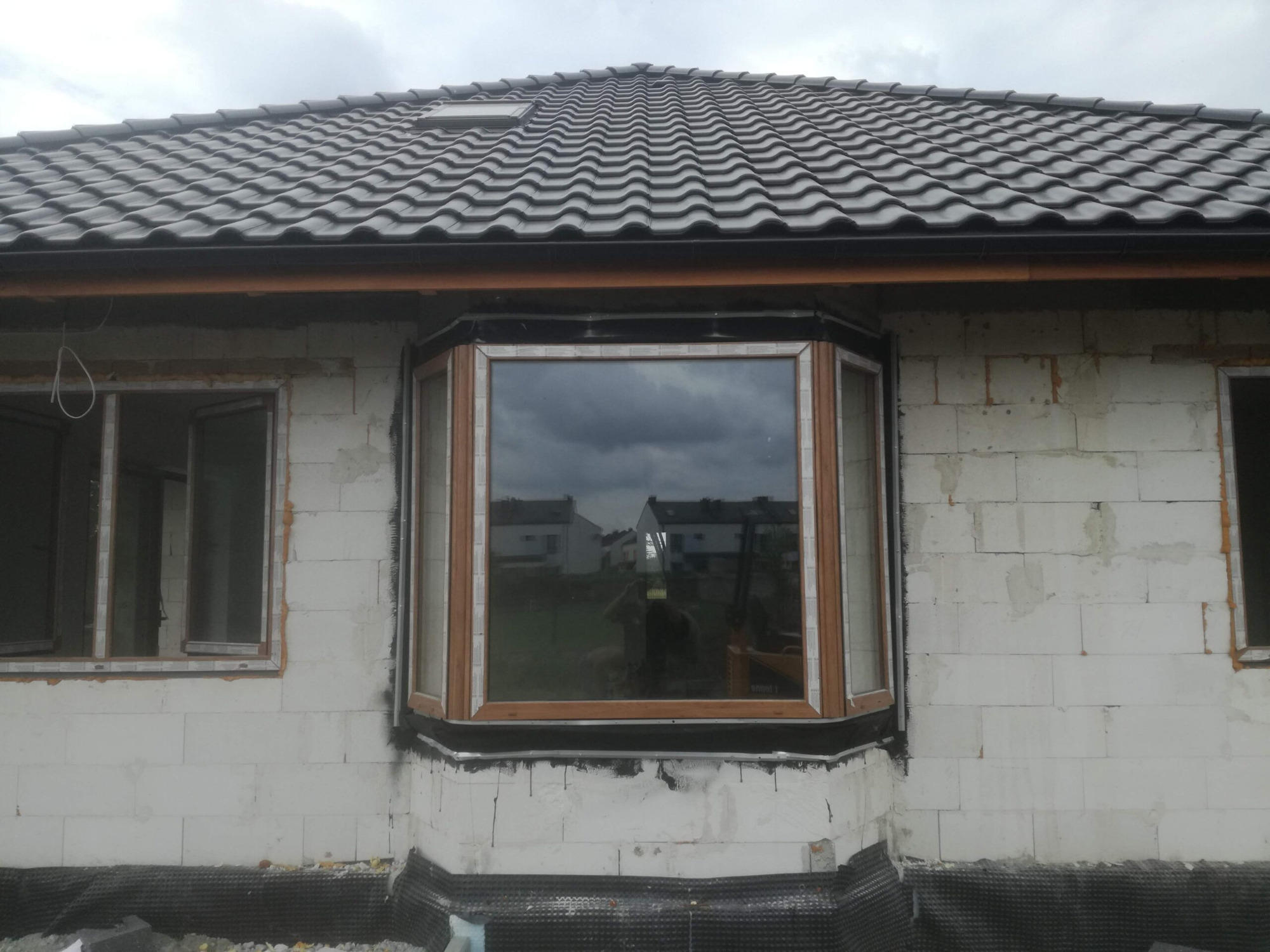 Wstawianie okna wykuszowego z drewnianą ramą w surowy mur z białych bloczków, widoczne przygotowania do montażu i fragment dachu z ciemnej dachówki.