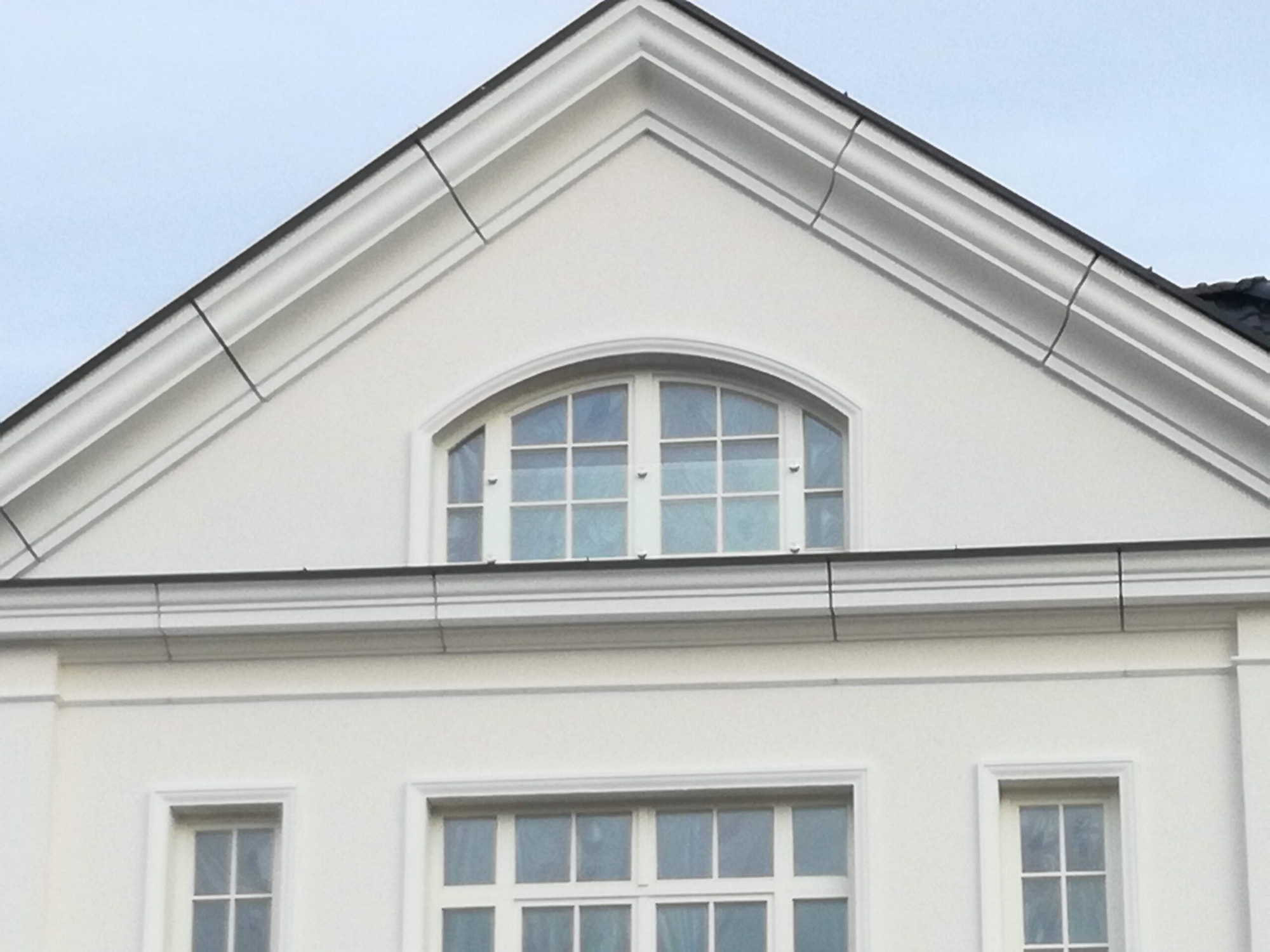 Fragment fasady budynku z białym tynkiem, widoczne trzy okna z białymi ramami: jedno łukowe na poddaszu i dwa prostokątne poniżej. Na dachu widoczne ciemne dachówki.