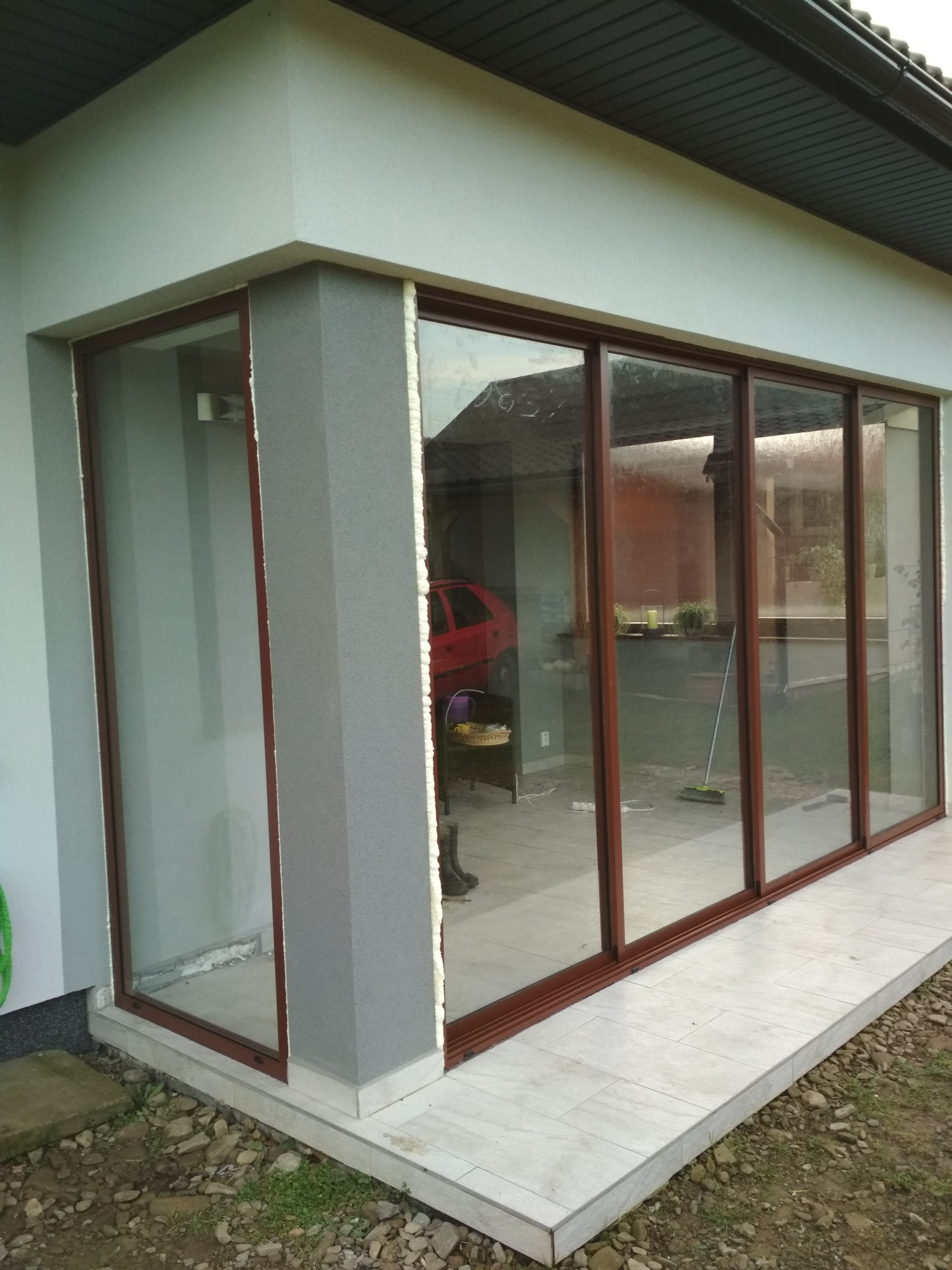 Zabudowa tarasu z przesuwnymi oknami z brązowymi ramami, widoczne pianowanie montażowe i odbicie czerwonego samochodu w szybach.