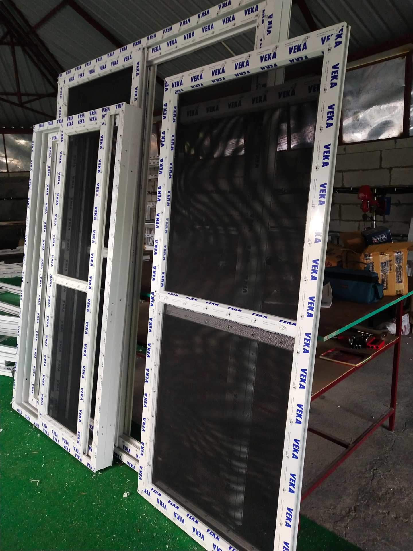 Białe ramy okienne PCV z siatkami moskitierowymi, ułożone w magazynie, widoczne napisy 'VEKA' na profilach.