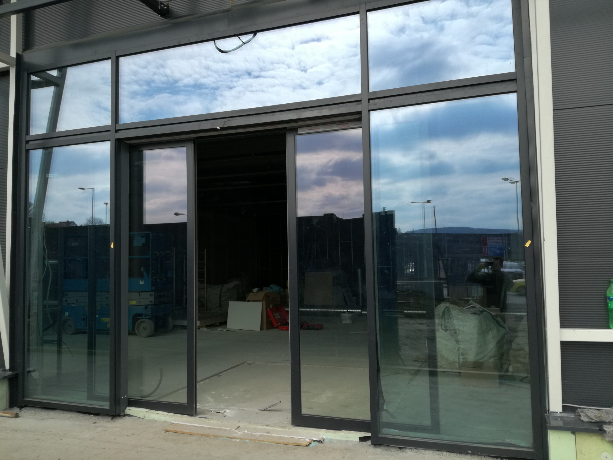 Nowoczesne, przesuwne, aluminiowe drzwi tarasowe w kolorze grafitowym, z widocznym odbiciem nieba i chmur w szybach, zamontowane w obiekcie w trakcie budowy.