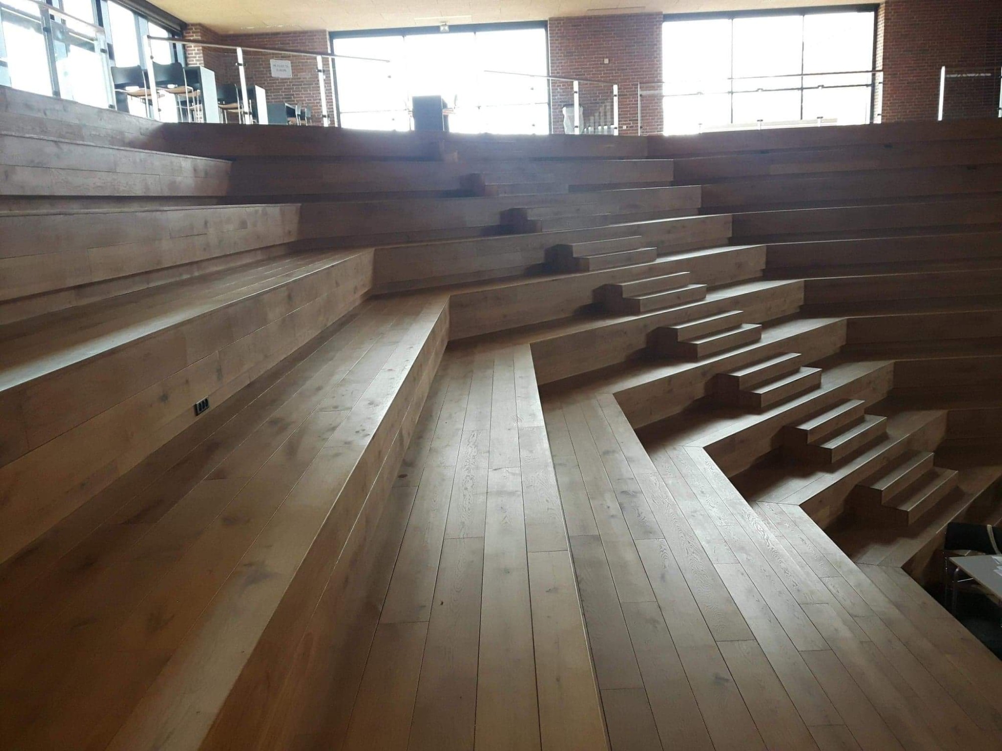 Amfiteatralne schody z jasnego drewna, przypominające parkiet, w jasnym wnętrzu z ceglaną ścianą i dużymi oknami.
