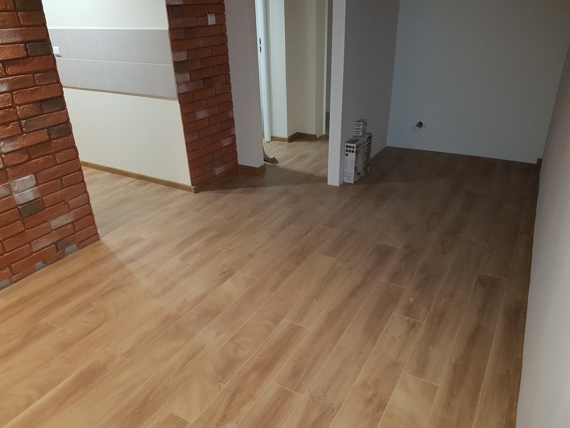 Jasne panele podłogowe imitujące drewno, ułożone w nowym mieszkaniu, widoczne fragmenty ceglanej ściany i białe ściany w tle.