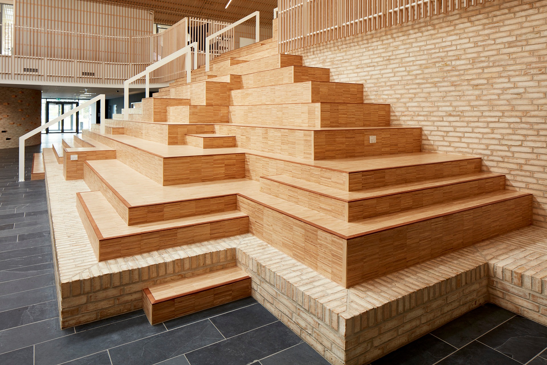 Nowoczesne schody z drewnianych paneli na ceglanej podstawie, prowadzące do wyższej części pomieszczenia z drewnianymi balustradami i ciemną podłogą z płytek.