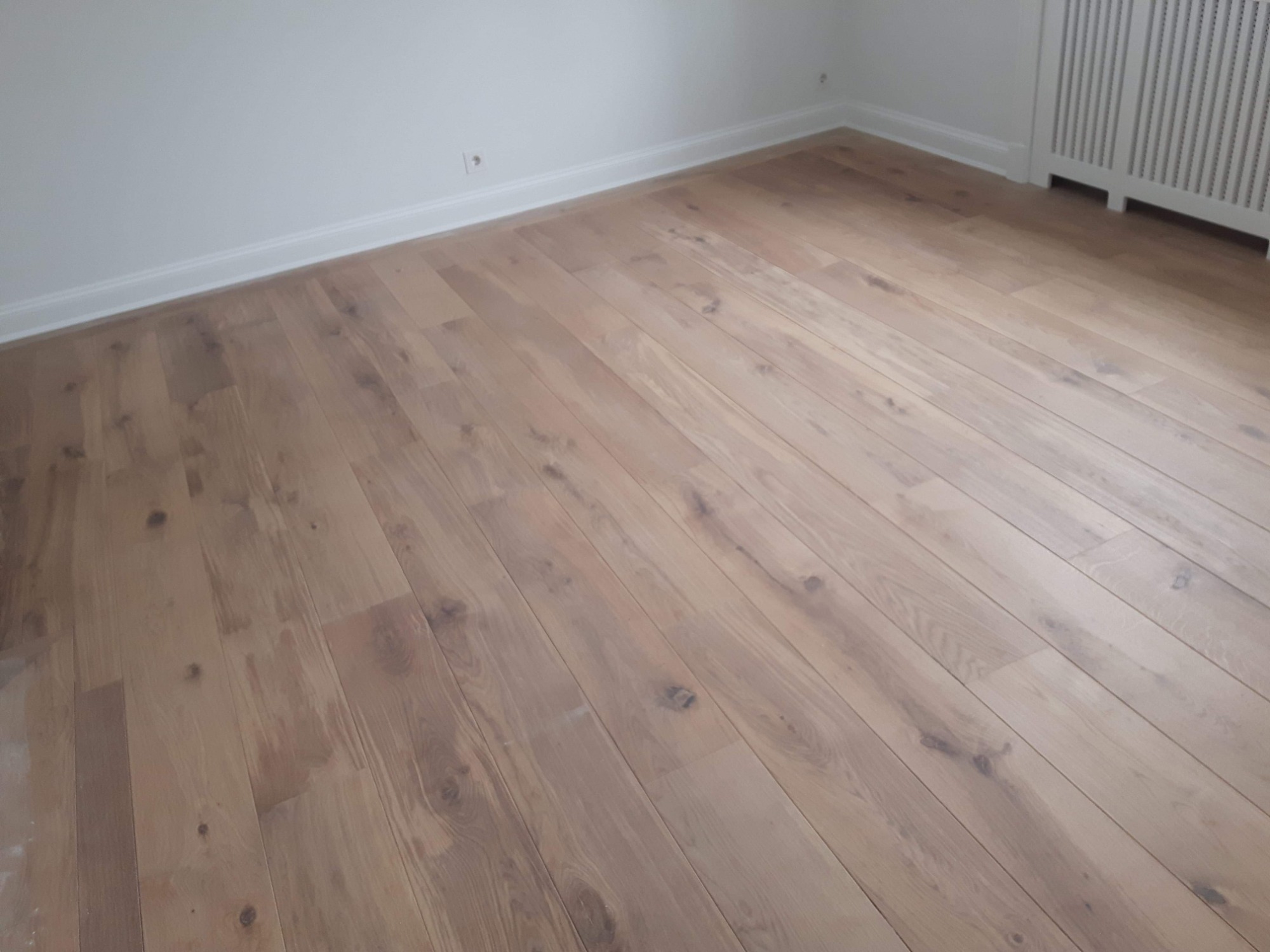 Jasny, drewniany parkiet ułożony w nowym, minimalistycznym wnętrzu, widok na grzejnik i listwy przypodłogowe