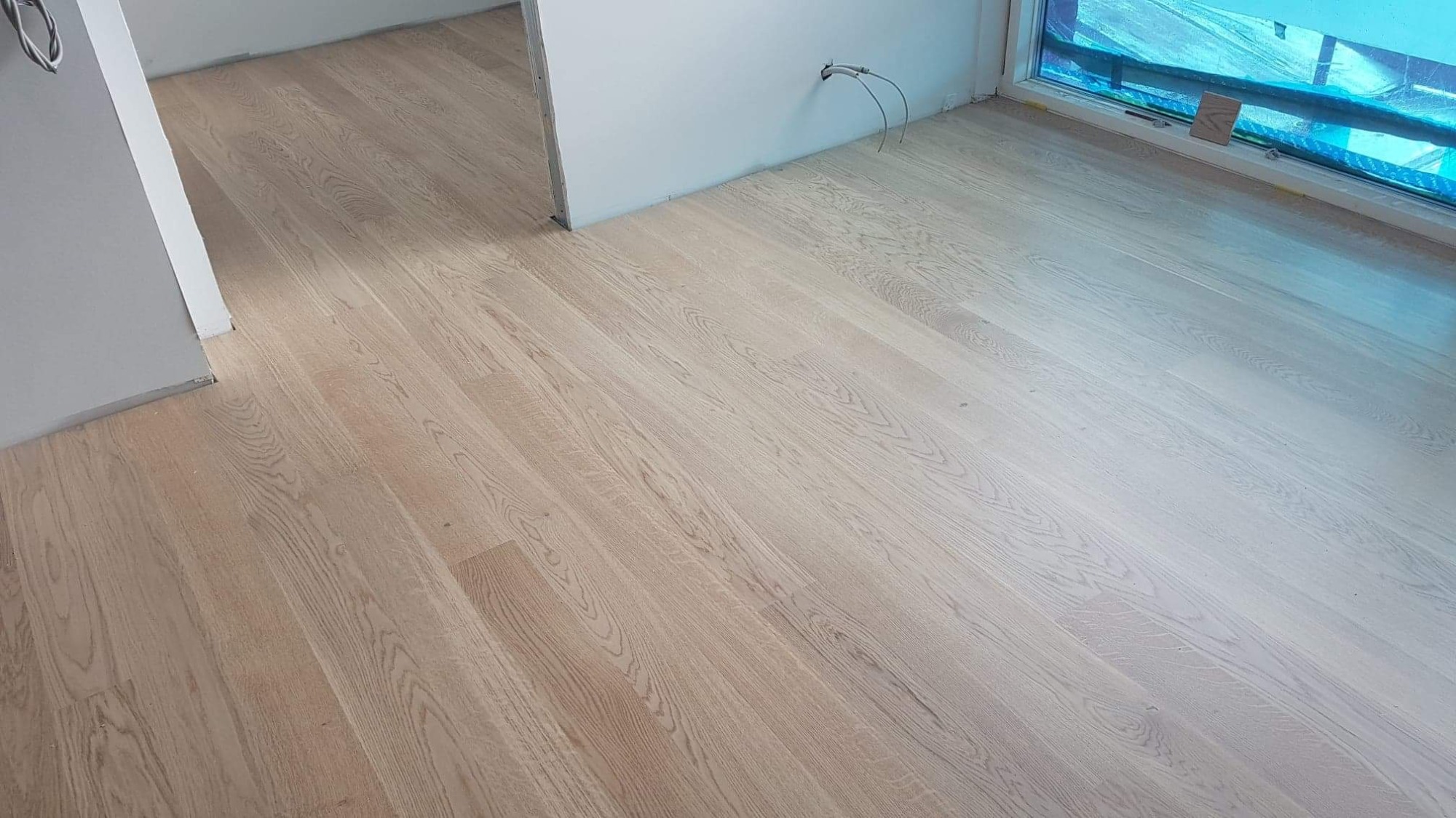 Jasny, lakierowany parkiet ułożony w nowym pomieszczeniu z widocznymi przewodami wystającymi ze ściany i oknem zabezpieczonym folią malarską.