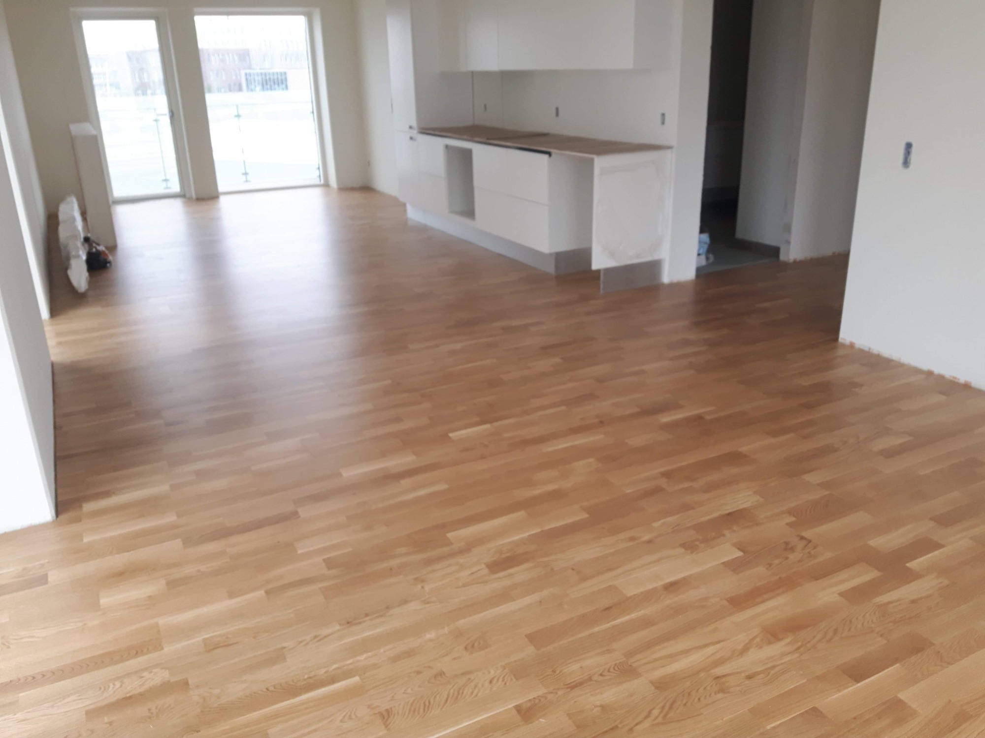 Jasny, nowo ułożony parkiet w przestronnym, minimalistycznym wnętrzu z białymi ścianami i zabudową kuchenną. Widoczne duże okna z widokiem na zewnątrz.