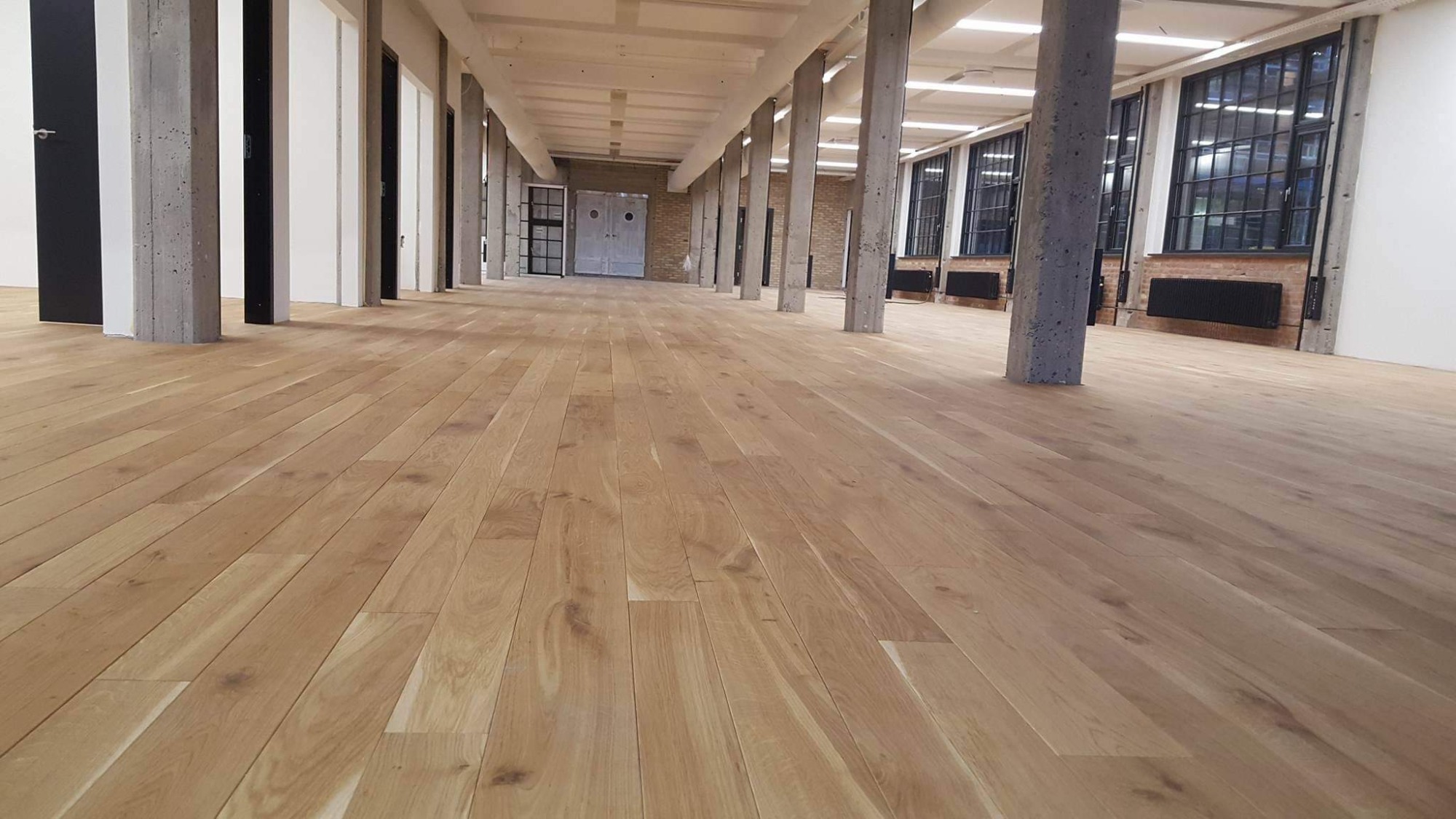 Jasna, drewniana podłoga ułożona w przestronnym, industrialnym wnętrzu z betonowymi kolumnami i dużymi oknami w czarnych ramach.