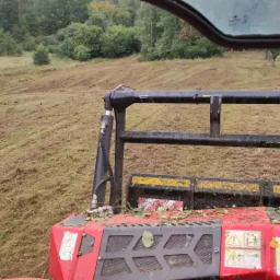 Widok z kabiny czerwonej maszyny rolniczej na świeżo skoszone pole trawy, z widocznymi śladami przejazdu i pozostałościami roślinności.