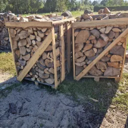 Dwa drewniane pojemniki na paletach wypełnione pociętymi kawałkami drewna, ustawione na piaszczystym podłożu z widoczną zielenią w tle.