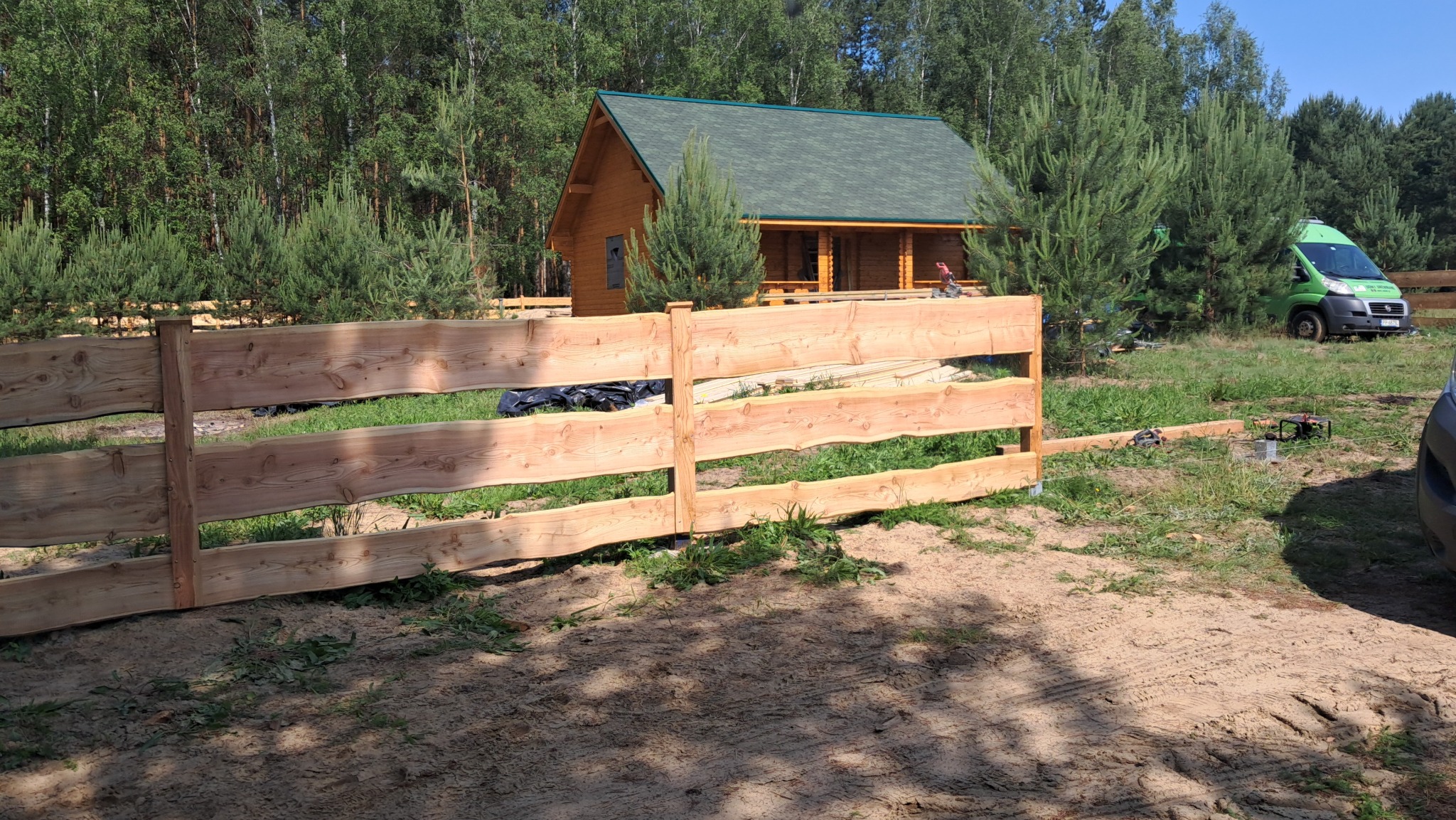 Naturalne ogrodzenie z surowego drewna przed domem z bali, z zielonym dachem. W tle las i zielony bus. Widoczne narzędzia sugerują montaż w toku.